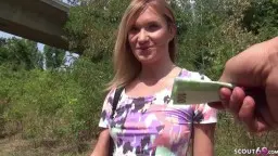 Junge blonde Frau mit frechem Lächeln steht in buntem Top auf sonniger Wiese unter Autobahnbrücke