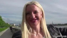 Strahlende blonde Russin mit langen Haaren spricht lächelnd in die Kamera vor Hochhaus und grünen Bäumen