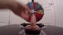 Nahaufnahme eines steifen Penis in transparenter Quetschvorrichtung, während eine Hand das bunte Schmerzensrad dreht