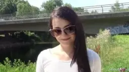 Brünette Milf in engen Shorts und weißem Crop-Top steht lasziv vor einer Brücke im Grünen