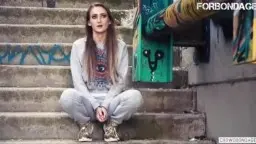 Verängstigte junge Frau mit langen braunen Haaren und traurigem Blick in grauem Pullover vor grüner Graffiti-Wand
