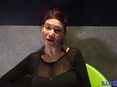 Reife rothaarige Frau mit großen Ohrringen und schwarzem Netzoberteil sitzt lasziv auf grünem Sessel und leckt sich lustvoll über die Lippen.
