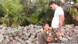 Junger Mann in Camouflage kniet vor großem Chef in weißem Shirt zwischen Steinmauer und Palmen
