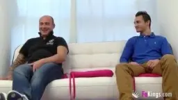 Ainara im kurzen Rock und roten Overknee-Stiefeln sitzt lasziv im schwarzen Designersessel neben einem tätowierten Mann auf weißem Sofa.