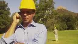 Bauarbeiter mit gelbem Helm telefoniert auf sonniger Wiese mit zweitem Arbeiter im Hintergrund