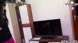Schwarzhaarige Frau im schwarzen Top und pinker Hose steht neben einem großen Fernseher im Wohnzimmer