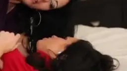 Zwei junge Dunkelhaarige im engen violetten und roten Top sitzen nah beieinander auf dem Bett und schauen sich voller Verlangen an