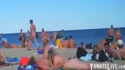 Nackte Frau liegt auf Decke am vollen Strand und wird von mehreren Männern umringt während sie oral verwöhnt wird
