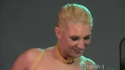 Blonde Frau mit Lederhalsband steht lächelnd neben einem schwarzen BDSM-Stuhl, während ein Mann in weißem Hemd ihn vorbereitet